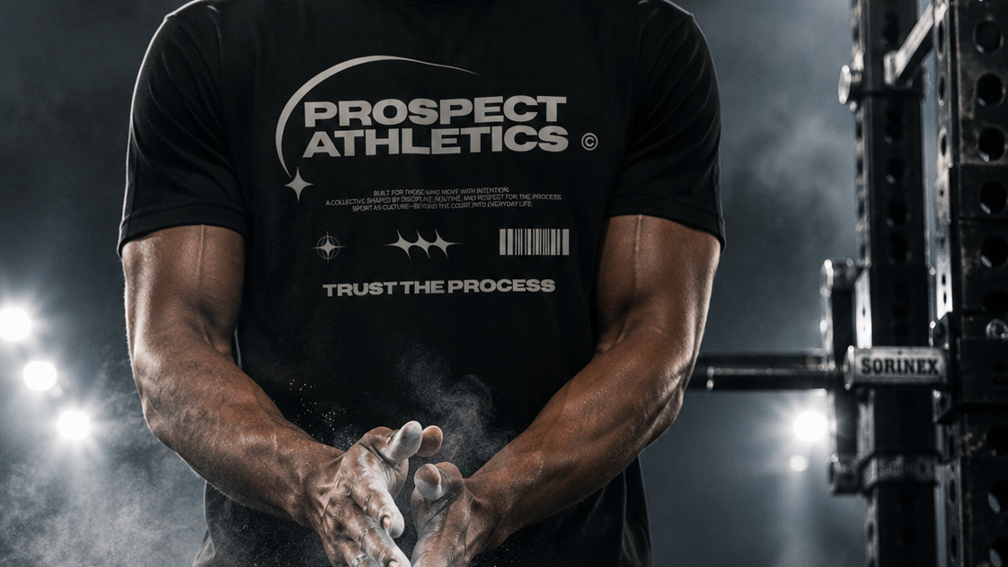 A muscular man in a gym wearing a black Prospect Athletics t-shirt and shorts, rubbing chalk on his hands in preparation for lifting. The background features a dark power rack and bright gym lights.
