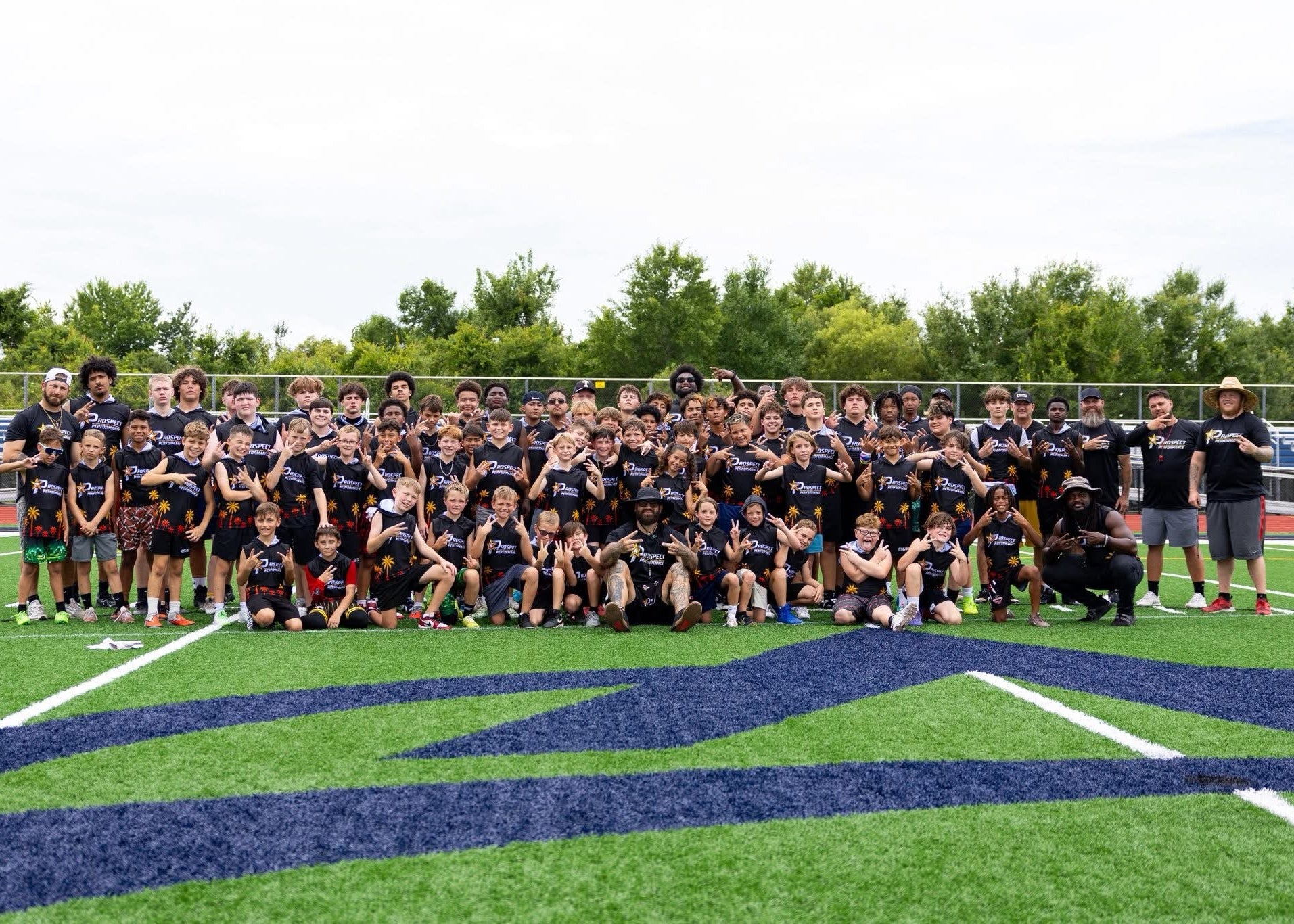 A large group of young athletes and coaches posing for a team photo on an outdoor football field. The children are wearing matching black athletic t-shirts with a white logo, while adults wear casual team apparel.